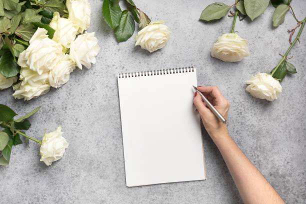 istock Female hands writing in notebook and bouquet white roses on grey. Top view with copy space. 1201610744