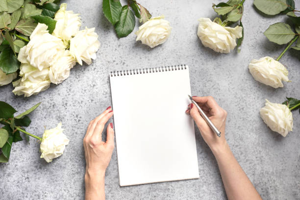 istock Female hands writing in notebook and bouquet white roses on grey. Top view with copy space. 1201610805