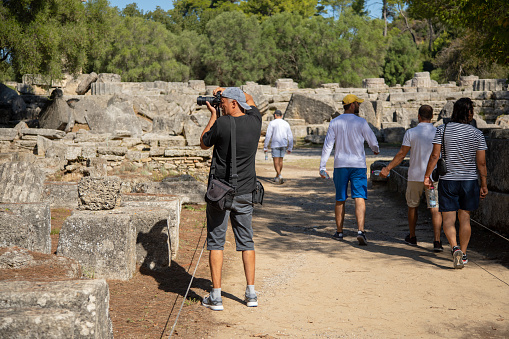 Man photographer taking a photo of columns and remaining of temple in archaeology site while visiting ancient Olympia, Peloponnese, Greece. Group of tourists passing by him.