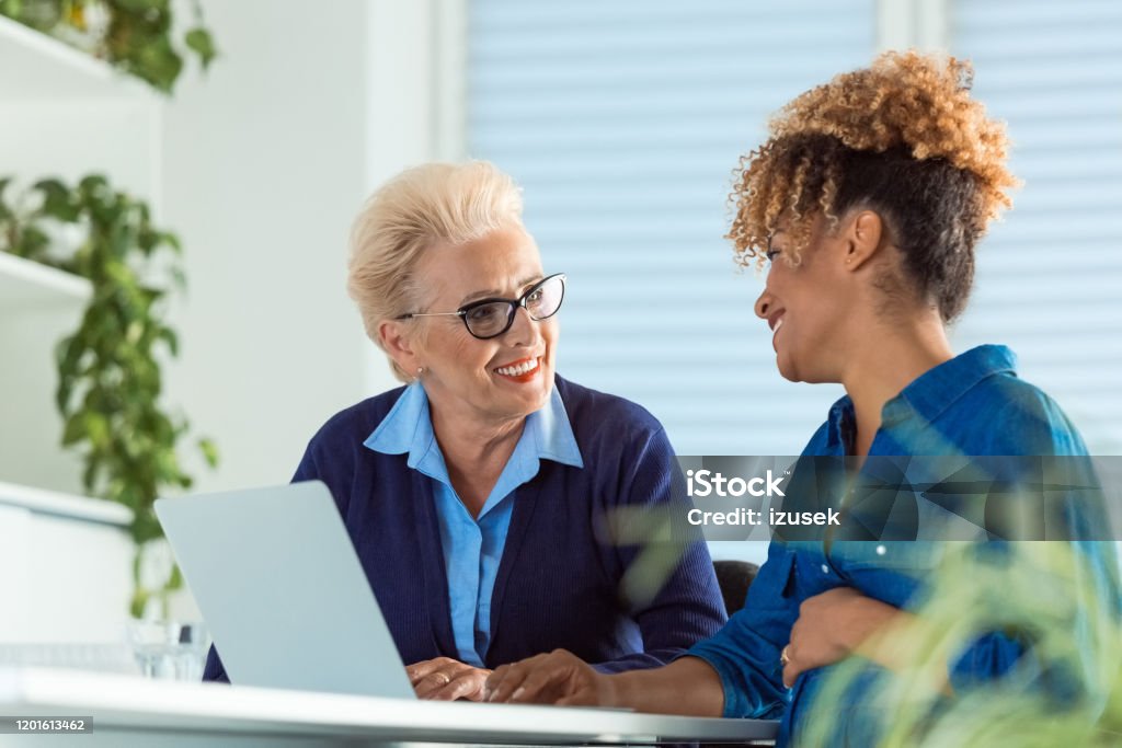 Pregnant executive looking at elderly role model Smiling female executives talking while using laptop. Pregnant businesswoman looking at elderly role model in meeting. They are discussing at desk in office. 35-39 Years Stock Photo