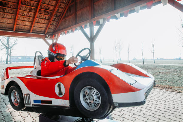 Young boy in a helmet and goggles sitting in a red and white toy racing car Boy dressed in red winter clothes is holding a steering wheel of a model toy car. kid toy car stock pictures, royalty-free photos & images