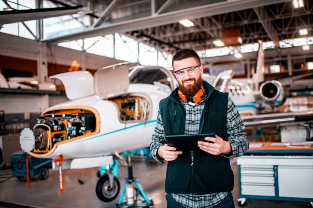 istock Aircraft mechanic in a hangar smiling and looking at a camera while holding a tablet and repairing and maintaining an airplane 1201618936