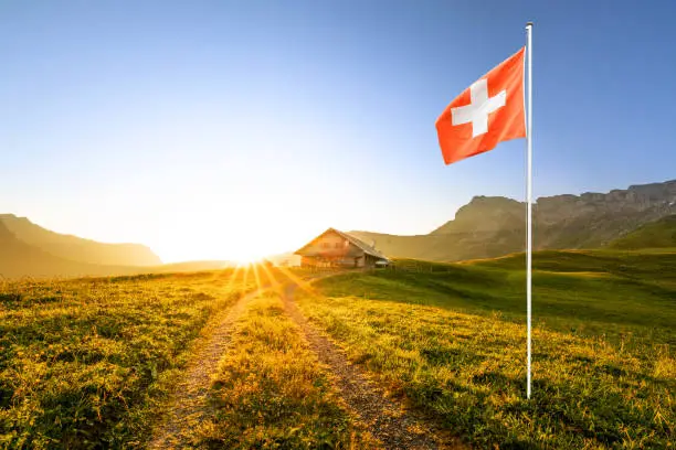 small road leading to a swiss chalet or farm in a mountain landscape at sunrise with sun star and a waving swiss flag in the foreground