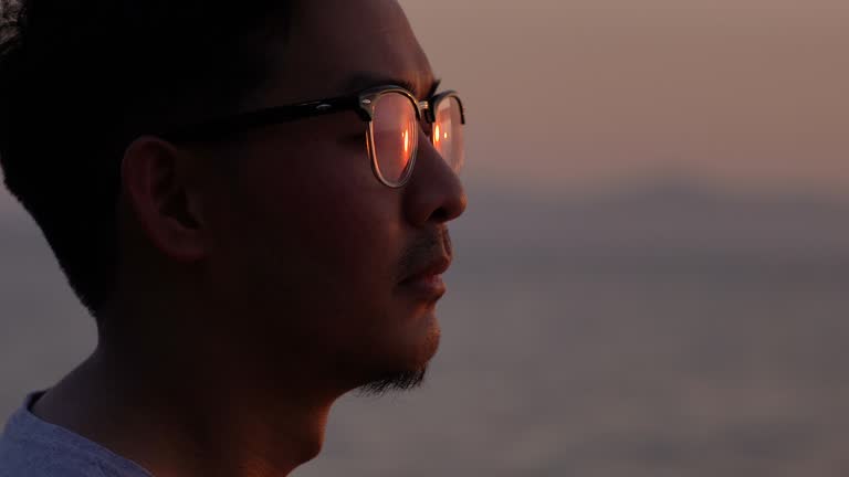 Close-up face of Asian man in eyeglasses looking at sunset with sunlight reflection