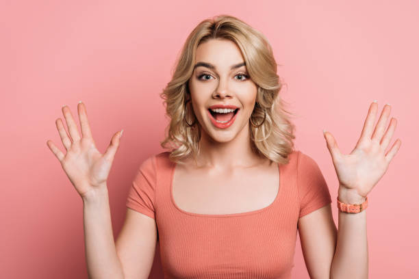 istock cheerful girl waving hands while smiling at camera on pink background 1201624852