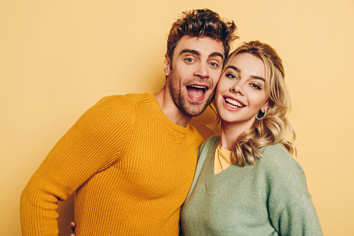 young, cheerful couple hugging while smiling at camera on yellow background