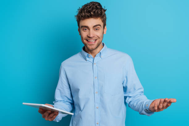 istock cheerful young man standing with open arm while holding digital tablet and smiling at camera on blue background 1201624884