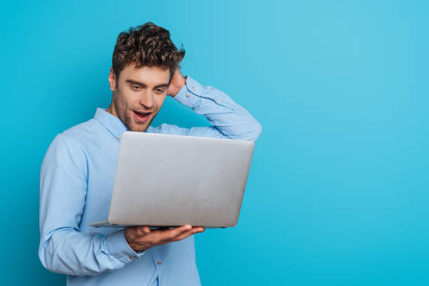 istock surprised young man touching head while looking at laptop on blue background 1201624887