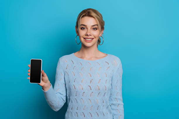 istock cheerful girl smiling at camera while showing smartphone with blank screen on blue background 1201624902