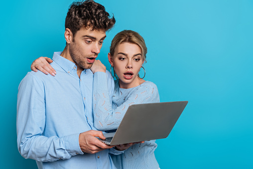 shocked girl hugging surprised boyfriend while looking at laptop together on blue background