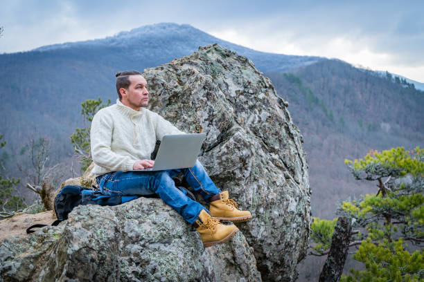 istock Hipster man working with notebook sitting on the rocky mountain on beautiful scenic clif background 1201629499