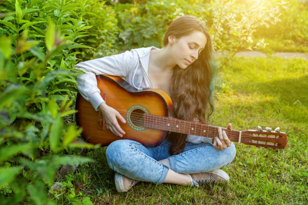 młoda hipsterka siedząca w trawie i grająca na gitarze na terenie parku lub ogrodu. nastolatka uczy się grać piosenkę i pisać muzykę - rock garden zdjęcia i obrazy z banku zdjęć