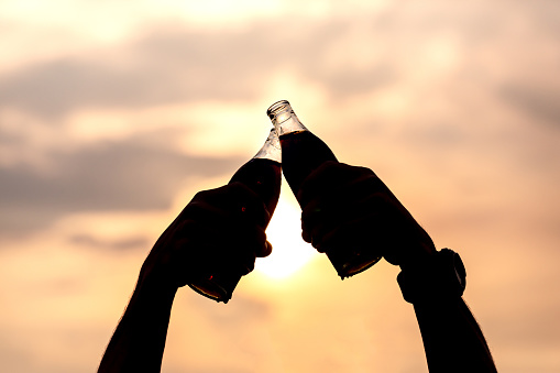 Hand of man clink soft drink bottles and raise on the sky in evening with sunlight.Silhouettes in the light of sunset.celebration and refreshment energy concept.