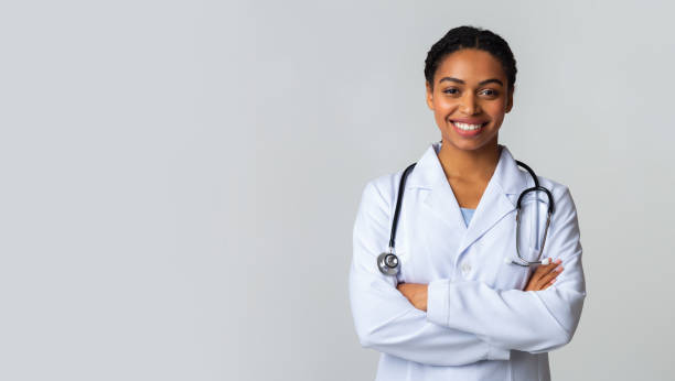 docteur féminin noir de sourire dans le manteau blanc posant avec les bras pliés - health care professional photos et images de collection