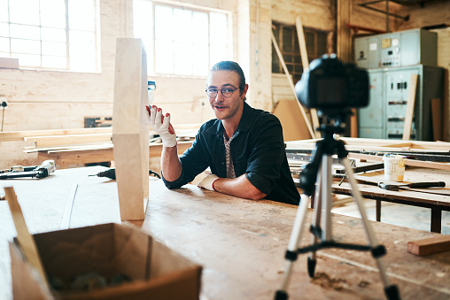 Shot of young carpenter recording a tutorial of him making a wooden masterpiece on a video camera inside his workshop