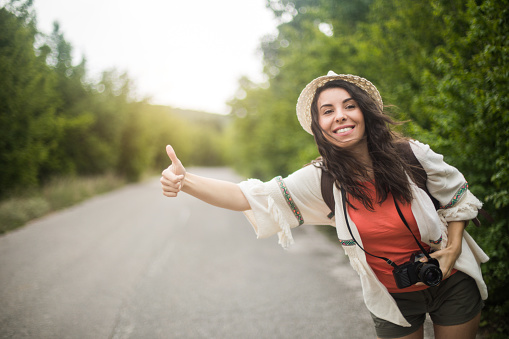 Beautiful young woman with backpack hitchhikes along road, keeps thumb raised, has delighted expression, has vacation trip. Pretty female waits for cars on asphalt road, explores new destination