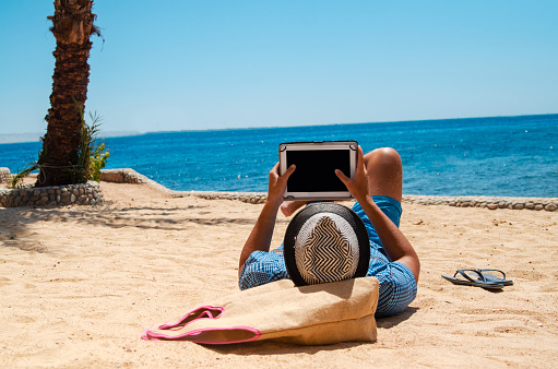 Young Man is reading on the beach with e-reader or surfing the net online with wifi connection.Relaxing with portable device.Copy space