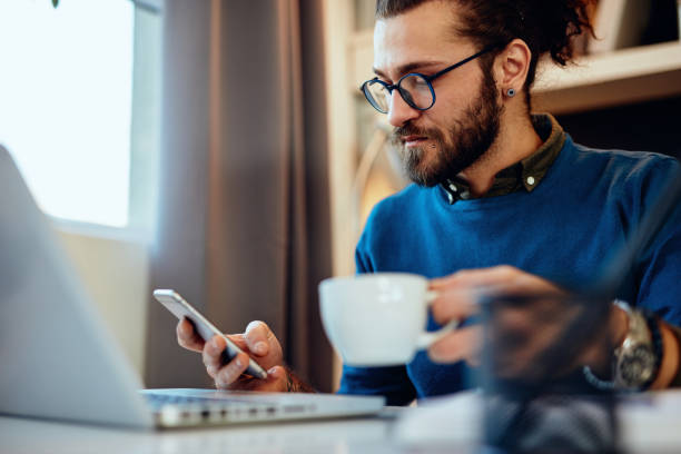 istock Young handsome bearded hipster sitting at his home office, holding cup of coffee and using smart phone. 1201668850