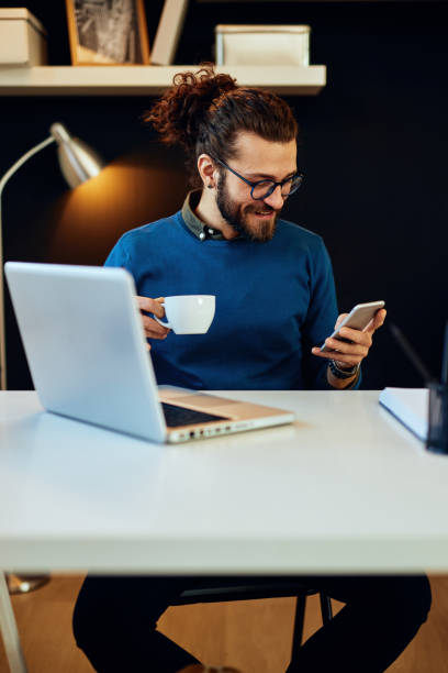 istock Young handsome bearded hipster sitting at his home office, holding cup of coffee and using smart phone. 1201668923
