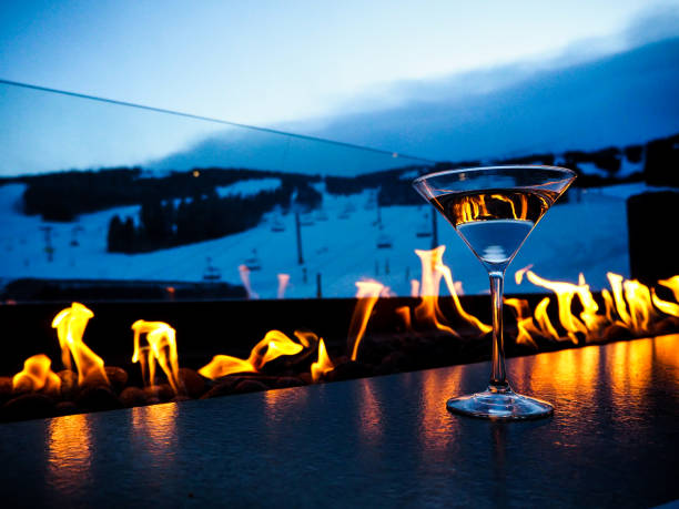 Apres Ski with a fire and a drink, in front of the slopes Relaxing in the evening after skiing with a martini in front of a fire table, with a view of the slopes, striking oranges, and contrasting blues, fire reflecting in the glass. apres ski stock pictures, royalty-free photos & images