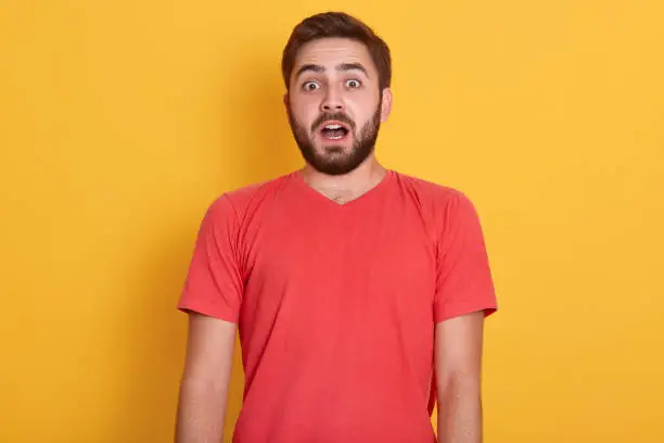Photo of Close up portrait of astonished bearde male, posing isolated over yellow background, handsome man standing with widely opened mouth, young guy wearing red casual t shirt. People emotions concept.