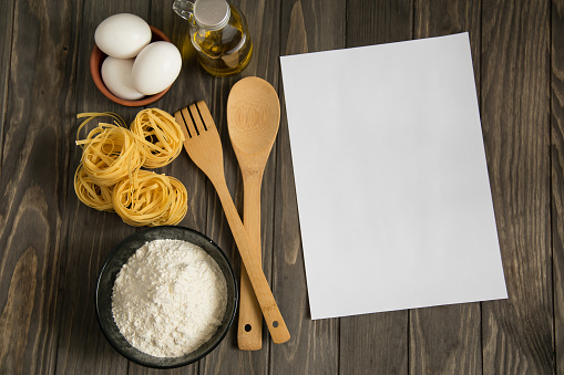 Recipe mockup or menu on wooden background bowl with flour, pasta, eggs, oil in a bottle, wooden spoon and fork, flat lay, copyspace for your text. Can be used to list diet foods.