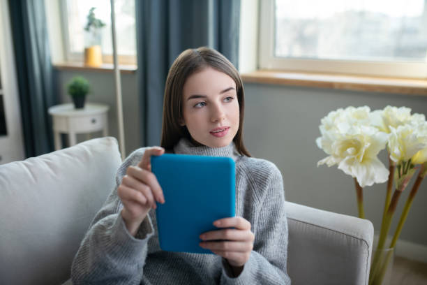 istock Dark-haired girl in a grey sweater with a tablet looking thoughtful 1201695312