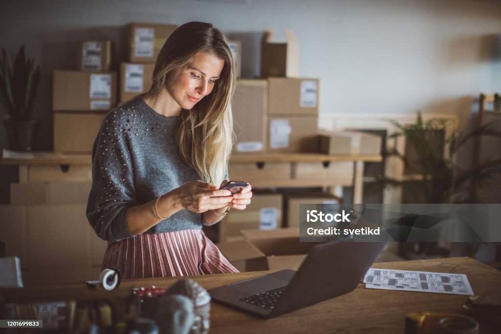 Organizing work in delivery business Working woman at online shop. She wearing casual clothing and using laptop and mobile phone for work organization Entrepreneur Stock Photo
