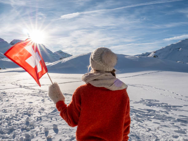 Cheerful young woman on mountain in winter holding Swiss flag in hand, snow winter wonderland
