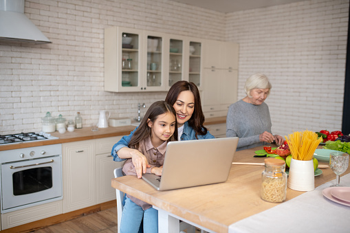In the kitchen. Young pretty woman with a joyful daughter working at a laptop sitting at a table, near a kind grandmother is preparing a salad.