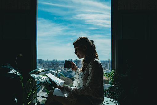 Silhouette of a young adult woman reading a notebook in front of a window