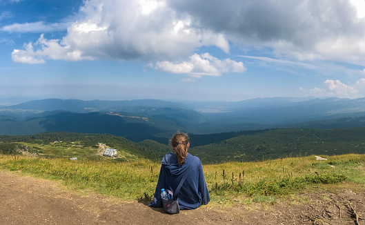 Blonde teen girl with ponytail sits by the path on the mountainside back in the frame in a blue hoodie, a bag, a bottle of water and looks at the breathtaking view from the top of the mountains