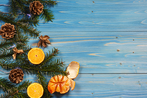Green fir branches with pine cones and sliced orange tangerines on the edge of a blue wooden table. Space for text