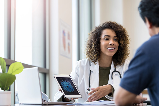 The mid adult female doctor holds the digital tablet that displays the x-ray and listens carefully to her unrecognizable patient.