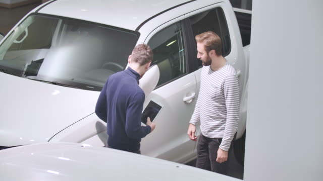 Top view of two Caucasian men standing next to the white pickup in car dealership. Male trader with tablet talking to customer in stripped sweater. Cinema 4k footage ProRes HQ.