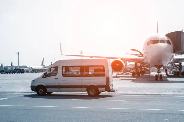 Photo of White VIP service van running on airport taxiway with big passenger airplane on background. Business class service at airport. Security intelligence agency hurrying at airfield