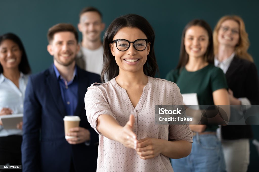 Mixed race woman sales manager stretch out hand greeting client Mixed race woman sales manager stretch out hand introduces herself greeting shake hands client smiling look at camera pose indoors with diverse teammates. HR, job interview, business etiquette concept Recruitment Stock Photo
