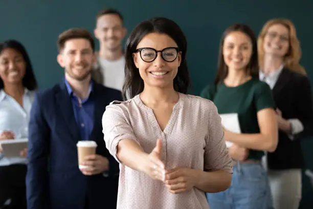 Mixed race woman sales manager stretch out hand introduces herself greeting shake hands client smiling look at camera pose indoors with diverse teammates. HR, job interview, business etiquette concept