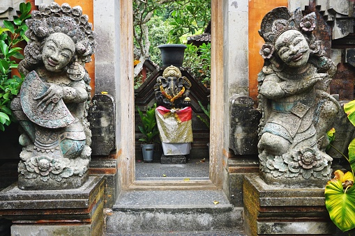 A vintage Ganesha statue can be seen through a doorway flanked by two carved stone figures with painted features near a temple in Bali Indonesia