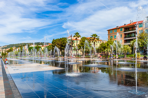 Tourists and locals in Promenade du Paillon of Nice, an urban area  with a large fountain, public park, wooden toys for children, lawns and benches to relax, palms, ficus trees and Mediterranean plants.