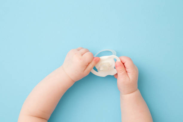 istock Infant hands holding white soother on light blue floor background. Pastel color. Closeup. First baby toy. Top down view. 1201823752