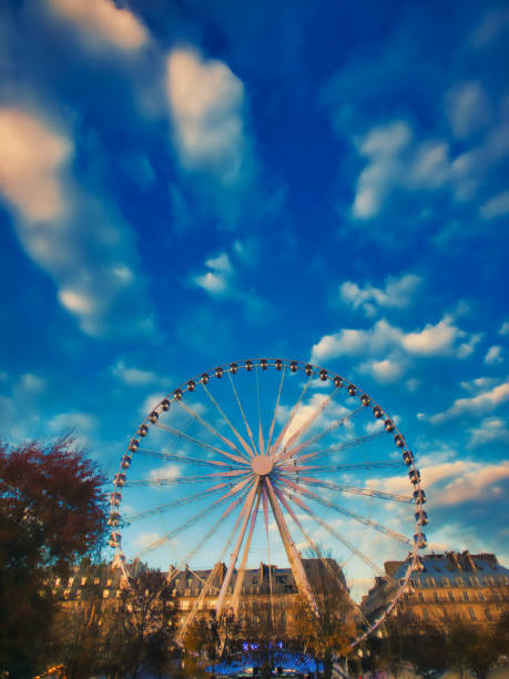 Paris, France - November 19, 2019 - View of the Ferris wheel from the Tuileries Garden in Paris lit by the sunset. In the foreground, the ferris wheel is visible behind trees in the garden. In the background several buildings on rue de Rivoli are visible