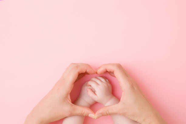 Heart shape created from young mother hands. Infant arms in middle. Pink pastel table background. Lovely emotional, sentimental moment. Closeup. Empty place for text, quote or sayings. Top down view.