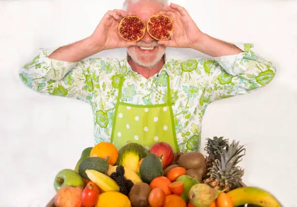 Photo of A cheerful senior man holds a pomegranate cut in half in front to his eyes. White wall on the background.  A basket full of healthy fresh fruit.