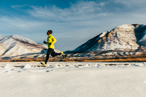 A middle aged male athlete goes for a cold, winter run in Utah, USA during a break in the snow storms. The sky is blue and the sun is shining. He is running to maintain his good mental and physical health and enjoying nature in the winter.