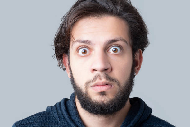 Close-up portrait of shocked young man looking at camera over gray background Close-up portrait of shocked young man looking at camera over gray background. Horizontal shot. confusion raised eyebrows human face men stock pictures, royalty-free photos & images