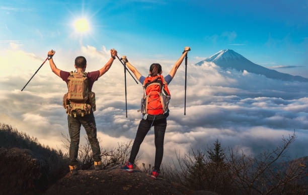 年輕的亞洲夫婦徒步旅行者爬上山尖山附近的富士。兩個背著背包的遊客在山頂享受日出。攀岩、説明和團隊工作理念 - 富士山 個照片及圖片檔