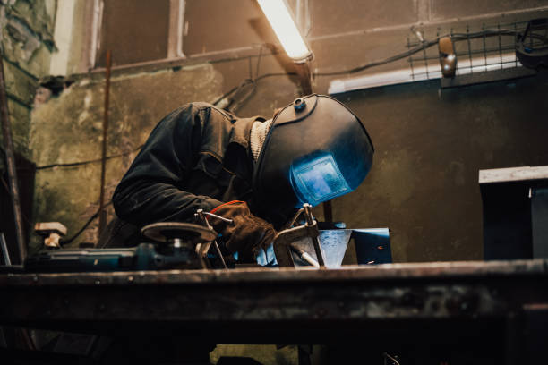 istock Worker in a welder mask works in a workshop for welding iron. Man makes iron products. Guy works with a welding machine using a mask to protect his eyes from dangerous rays 1201868825
