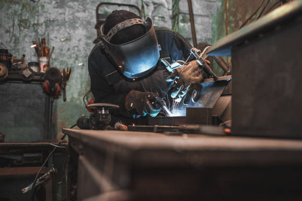 Worker in a welder mask works in a workshop for welding iron. Man makes iron products. Guy works with a welding machine using a mask to protect his eyes from dangerous rays