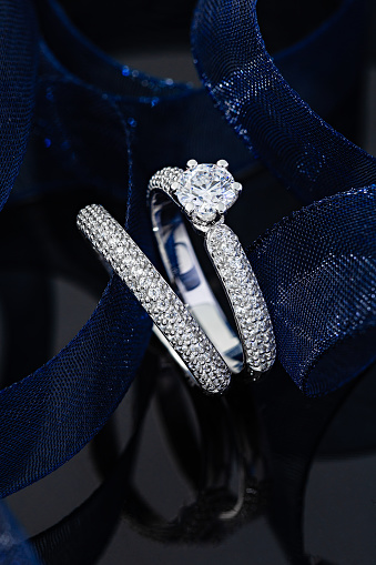 Wedding diamonds rings on black background with blue ribbon. Silver engagement rings with gemstone. Advertising jewelry still life